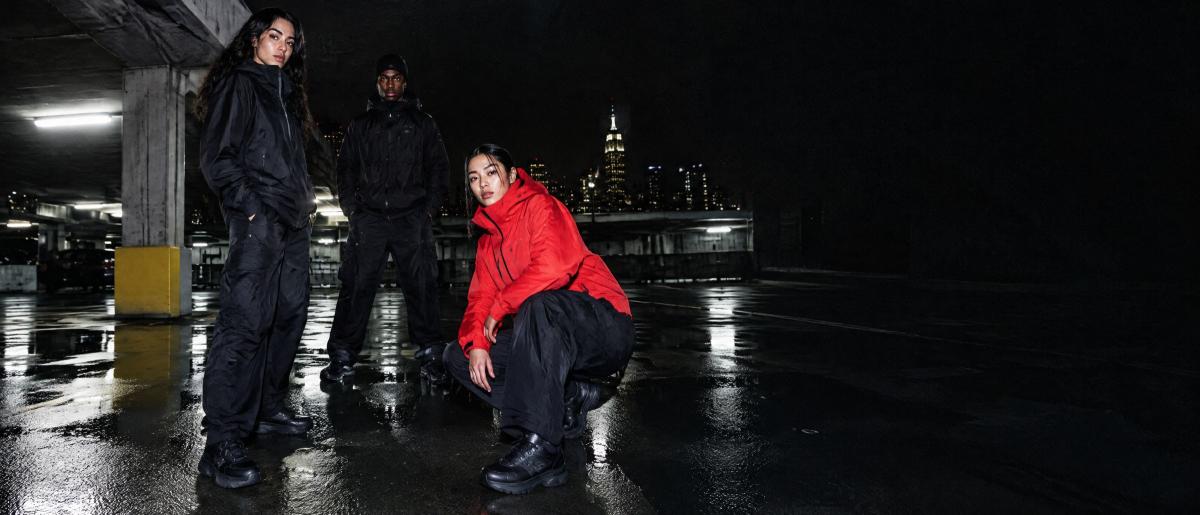 A woman in a red jacket crouching on a wet rooftop at night, the city behind