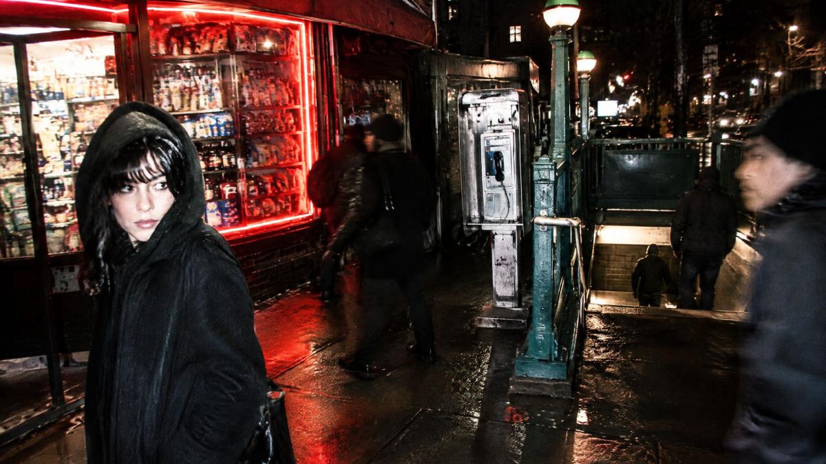 A woman in a black hood outside a lit corner store at night