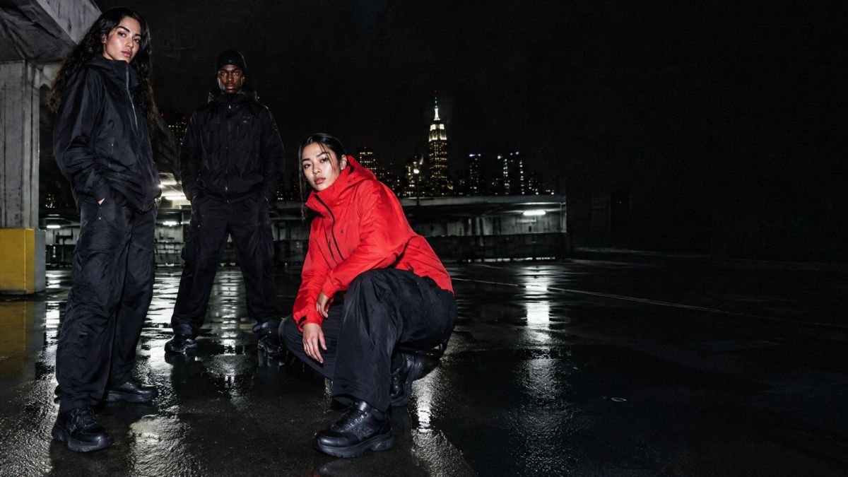 Three people in black and red jackets on a wet rooftop, the skyline behind