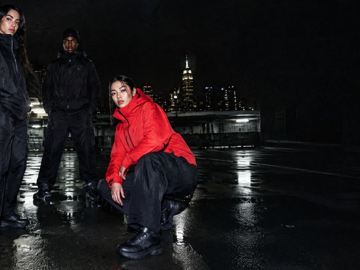 Three people in black and red jackets on a wet rooftop, the skyline behind