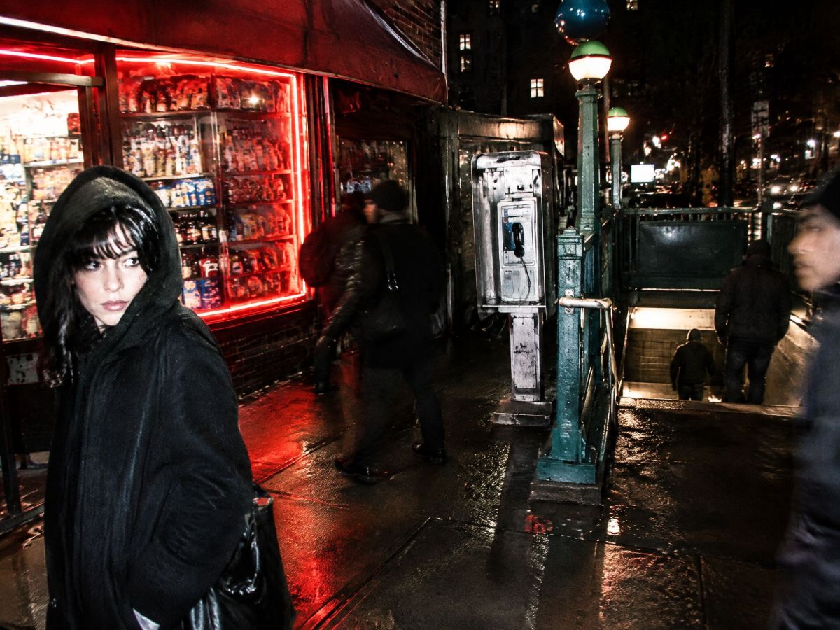A woman in a black hood outside a lit corner store at night