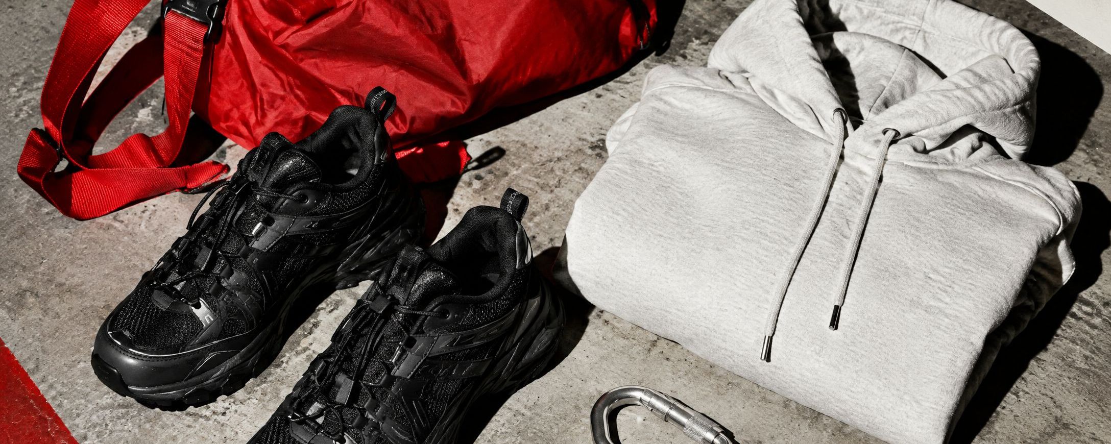 A red nylon bag, black runners, a gray hoodie and a carabiner on concrete