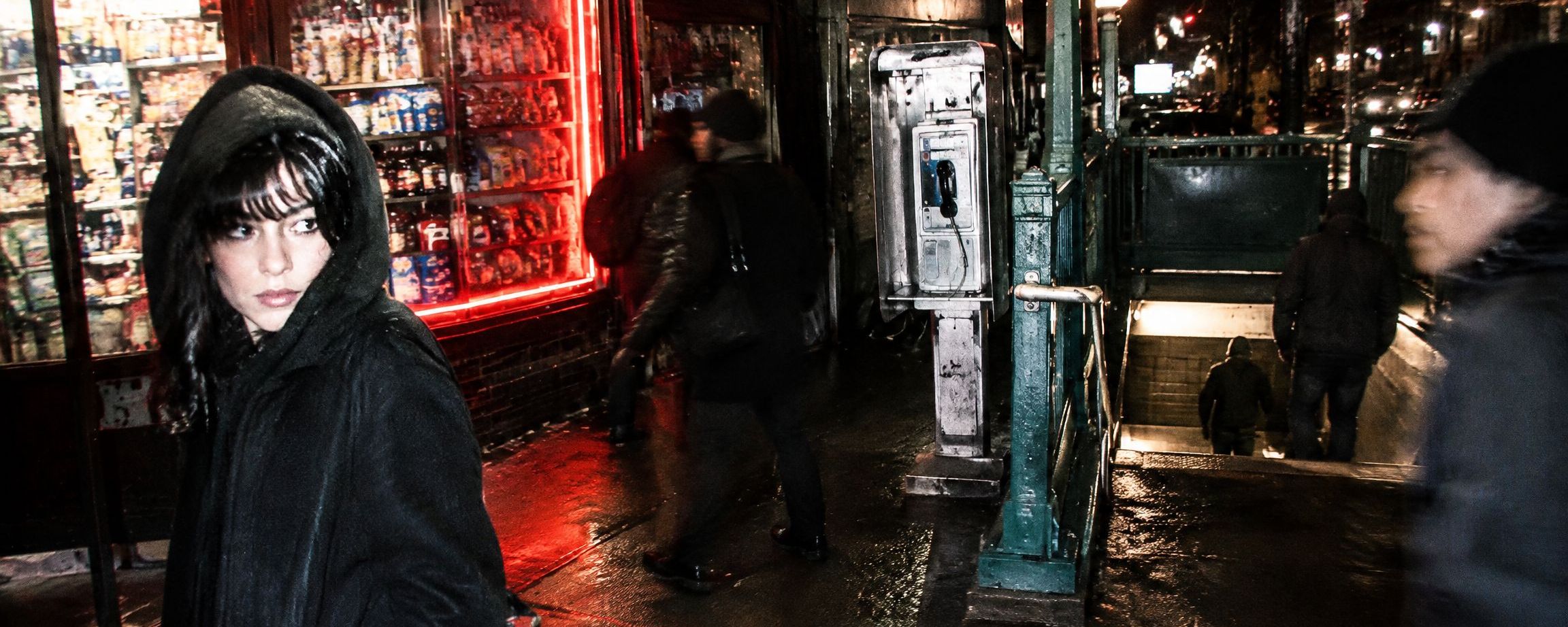 A woman in a black hood outside a lit corner store at night